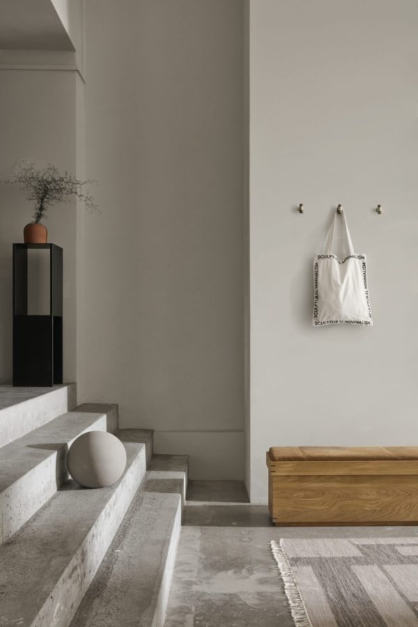 En minimalistisk entré med betontrin, en træbænk og en beige væg med en mulepose hængende. Til venstre holder et lækkert sort konsolbord en KRISTINA DAM - Dome Vase Ekstra Lille med grene. Et tekstureret tæppe ligger på gulvet.