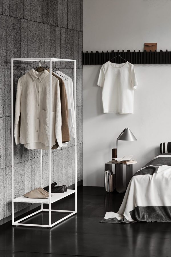 Et stilfuldt moderne soveværelse med et minimalistisk metaltøjstativ med et udvalg af skjorter og sko. Ved siden af fremhæver en pænt redt seng, en dekorativ lampe og nogle designmøblerbøger