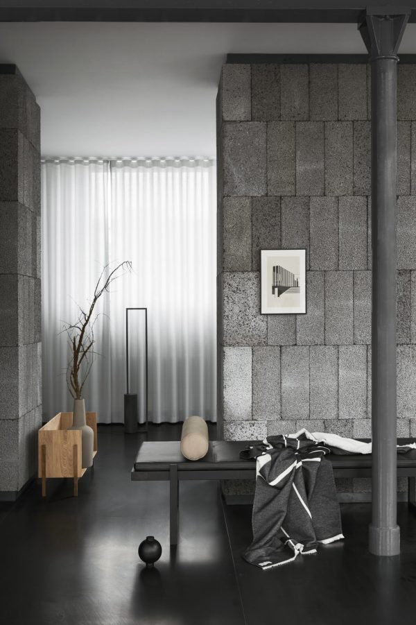 Moderne minimalistisk stue med grå stenvægge, en central metalsøjle, rene hvide gardiner, en træstol, en lav sort KRISTINA DAM - Minimal Daybed med et tæppe og dekorative genstande. neutrale toner dominerer rummet.