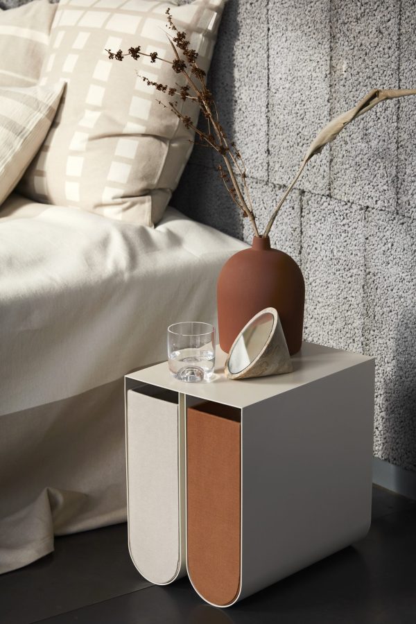 En moderne sengekantsopsætning med et hvidt og brunt buet natbord med en terracottavase med tørrede grene, et klart glas vand og en lille hvid skål. Naturligt lys oplyser scenen