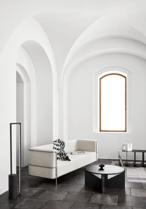 En minimalistisk stue med hvælvede hvide lofter, med en lysegrå sofa, et sort rundt sofabord og en slank, sort gulvlampe. Rummet har mørke klinkegulve og en lysende