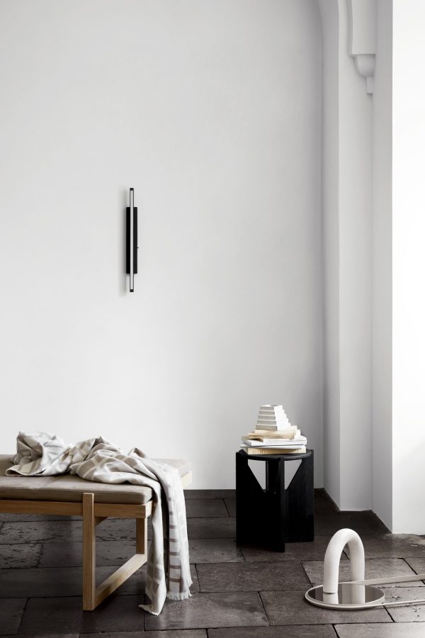 Minimalistisk værelse med buet hvid væg, træbænk med et draperet tæppe, stablede hvide bøger på en sort piedestal, rektangulær væglampet og en moderne designskulptur med sløjfe på gulvet.