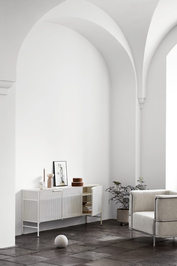 Et minimalistisk interiør med et hvidt skab med dekorationsgenstande, indrammede kunstværker og en potteplante, placeret under elegante buede lofter. En moderne sofa og spredte bøger fuldender den fredfyldte scene.