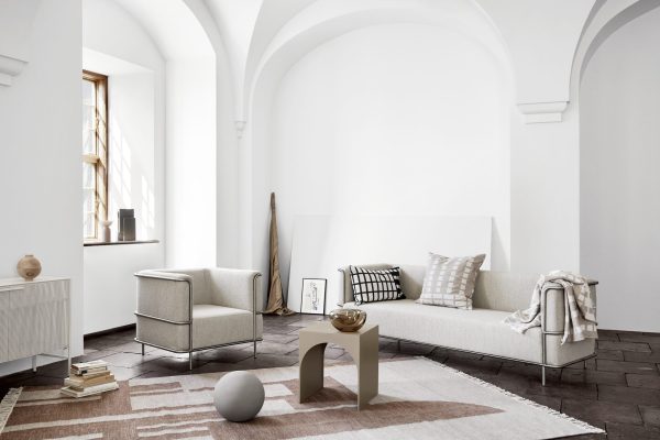 En minimalistisk og lys stue med hvide vægge, buede lofter, en beige sofa med puder, en hvid lænestol, et sofabord i træ og en KRISTINA DAM - Contemporary Pude på et mørkt klinkegulv.