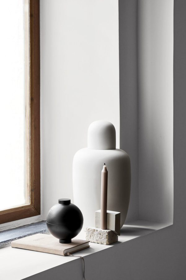 Minimalistisk stillebenskomposition på en vindueskant med en stor hvid vase, en mindre sort vase, en lysestage i sten med et beige lys og en åben bog. Sollys oplyser scenen