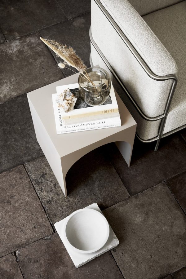 Et stilfuldt komponeret billede med et lille, hvidt sofabord ved siden af en moderne sofa på et mørkt klinkegulv. bordet rummer bøger, en glasskål med skaller og en tidsel med en KRISTINA DAM - Opal Vase Lille på gulvet ved siden af.