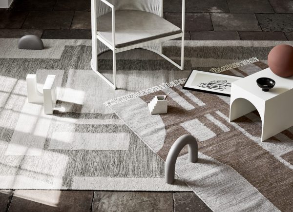 Et moderne minimalistisk værelse med et geometrisk mønstret tæppe, designmøbler inklusiv en hvid metalstol og abstrakt formede indretningsgenstande, herunder sfæriske og buede skulpturer på et klinkegulv.