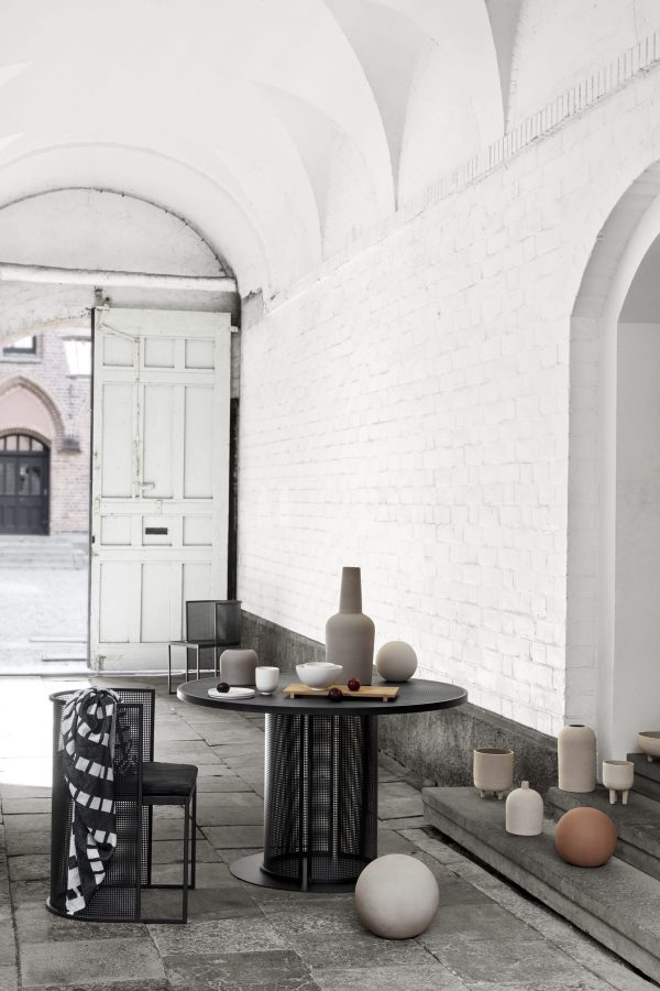 Et stilrent, minimalistisk interiør med sort rundt bord og diverse KRISTINA DAM - Dome Vase Ekstra Lille i neutrale toner. scenen er sat under buede hvide murstenshvælvinger, og et tekstureret kast er draperet over en stol, hvilket tilføjer varme.