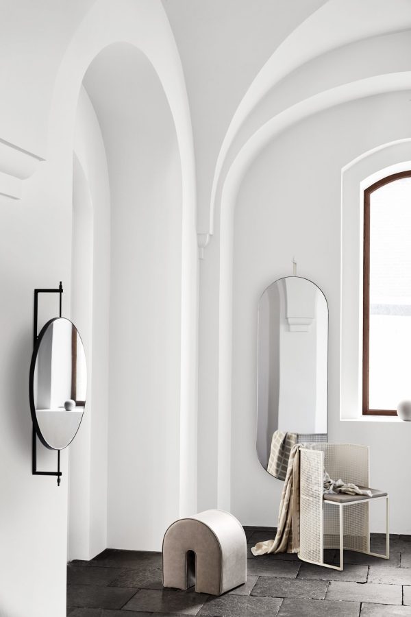 Et minimalistisk interiør med buede hvide vægge, buede døråbninger og et mørkt klinkegulv. Der er tre spejle på væggene, et rundt væghængt spejl og et designmøbler abstrakt