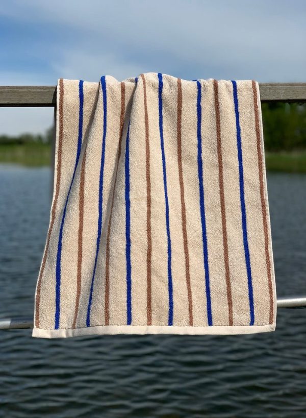 Et beige håndklæde med lodrette blå striber hængende på et metalgelænder af designermøbler med en sløret vand- og grøn baggrund.