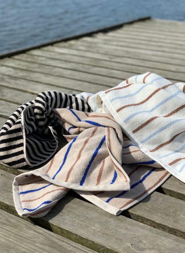 Et stribet håndklæde er afslappet draperet over en trædok ved siden af en fredfyldt vandmasse og indfanger en afslappet, solrig dag med et strejf af Tomorrows-design.