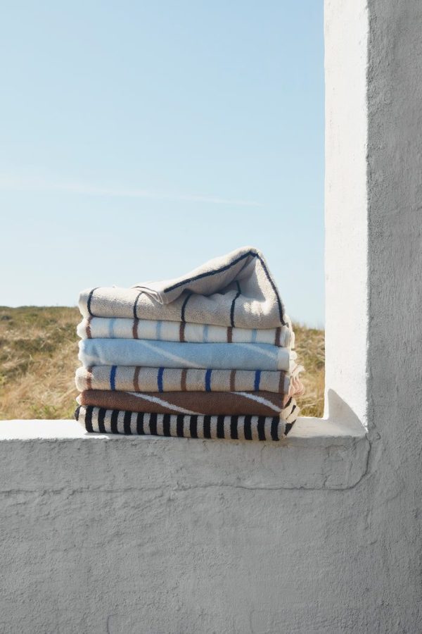 En stak foldede tæpper med forskellige stribede mønstre fra Tomorrowsdesign sidder på en hvid betonkant mod en klar himmel baggrund, med græsklædte klitter synlige i det fjerne.