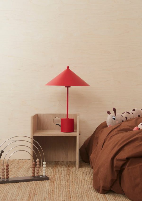 Et hyggeligt børneværelseshjørne med en rød lampe på et lille trænatbord ved siden af en seng med et brunt tæppe. Et plys giraflegetøj hviler på sengen og en møblerperle