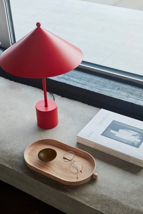 Rød bordlampe med Tomorrows-design hviler på en betonvindueskarm ved siden af en træbakke med glas og en dekorativ sten, nær en åben bog.
