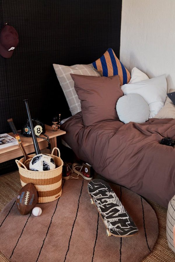 Et hyggeligt soveværelseshjørne med en brun seng med blå og stribede puder, et skateboard, en fodbold og designermøbler som en kurv med bøger ved siden af. En baseballkasket hænger på væggen.