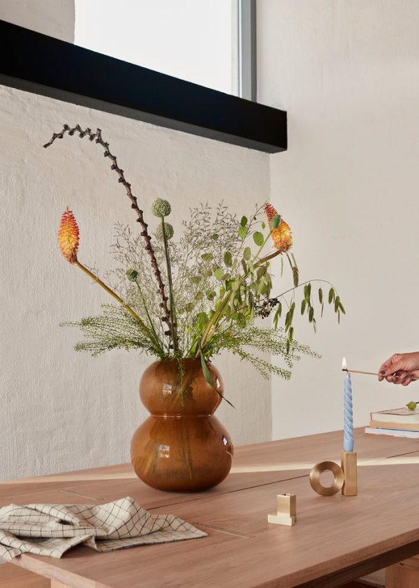 En elegant trævase med levende blomster på et enkelt spisebord. En hånd tænder et snoet blåt lys ved siden af dekorative messinggenstande og en foldet serviet, alt sammen arrangeret i et elegant Tomorrows-designinterval