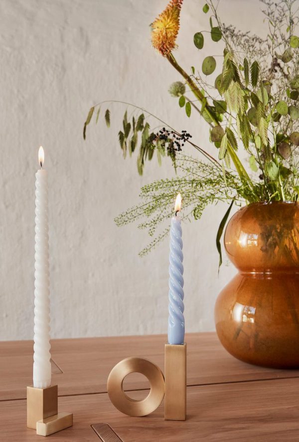 To tændte, snoede stearinlys i moderne guldholdere på et træbord, med i Tomorrowsdesign interiørkollektion, med en stor kobbervase indeholdende et frodigt arrangement af forskellige grønne planter og