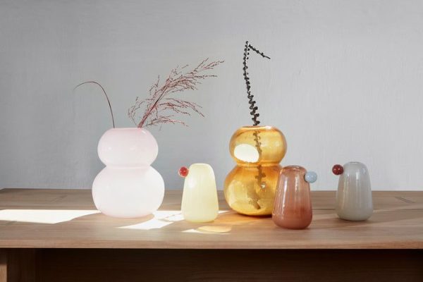 Fem unikke glasvaser med forskellige former og farver, der viser tørrede planter, er arrangeret på et træbord fra Tomorrowsdesign mod en almindelig væg oplyst af naturligt lys.