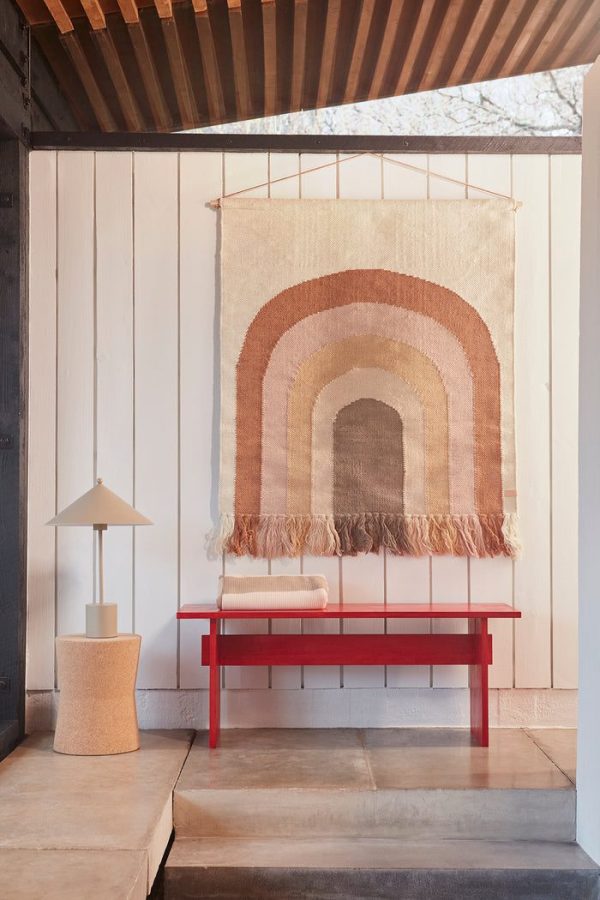 Et stilfuldt interiør med en rød OYOY - Kotai Bænk under et stort, flerfarvet vævet vægbeklædning. En cremelampe på et tekstureret sidebord komplementerer den enkle, elegante indretning.