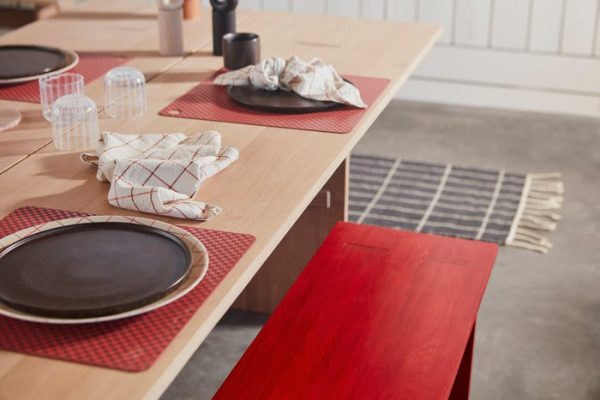 Et moderne spisebord sat med sorte tallerkener på røde dækkeservietter, parret med klare glas og hvidmønstrede servietter. OYOY - Kotai Bænk, der komplementerer trægulvet, er synligt.