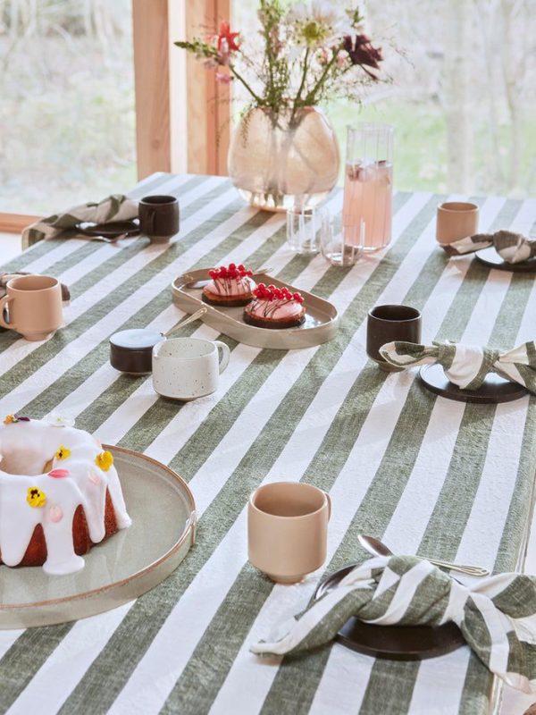 Et hyggeligt spisebordssæt med desserter inklusive en bundt-kage og tærter, keramiske krus og tallerkner på en OYOY - Striped Dug 260x140 dug, med vinduesudsigt over træer i baggrunden.