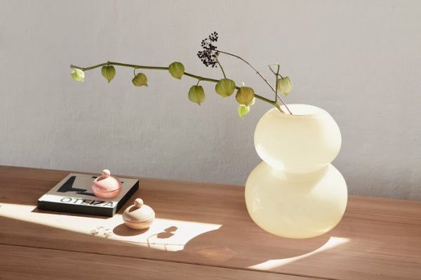 En moderne skulpturel bordlampe fra Tomorrowsdesign, med stablede sfæriske sektioner, oplyst på et træbord ved siden af en bog og små dekorative genstande, der kaster blødt lys på en hængende grøn plante med lillebitte