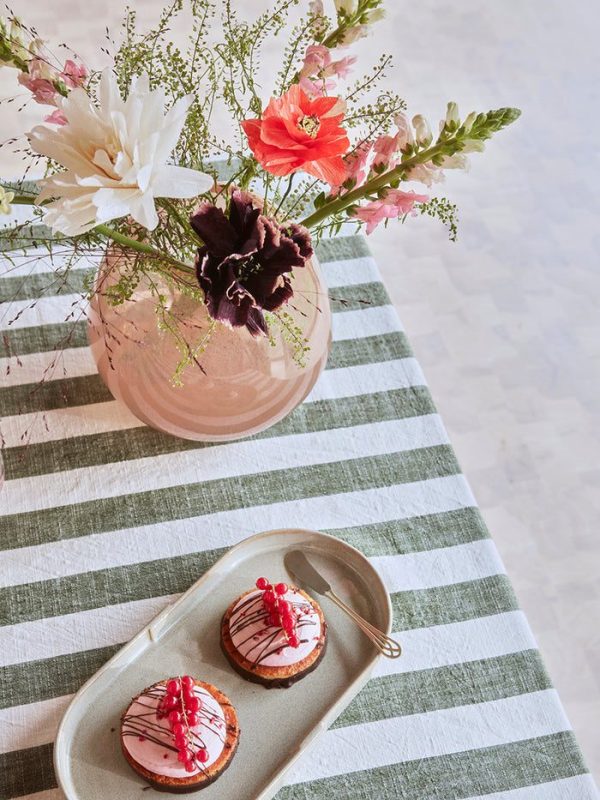 En række blomster i bløde nuancer arrangeret i en designer lyserød vase på et bord med en grå og hvid stribet dug. Ved siden af vasen står en rektangulær bakke med to lyserøde frostede småkager