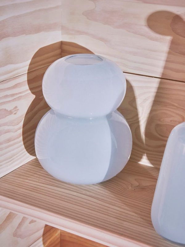 En vase af matteret glas med et afrundet, dobbelt-pære-design sidder på et træpanelgulv med blødt, plettet sollys, der kaster blide skygger omkring sig.
