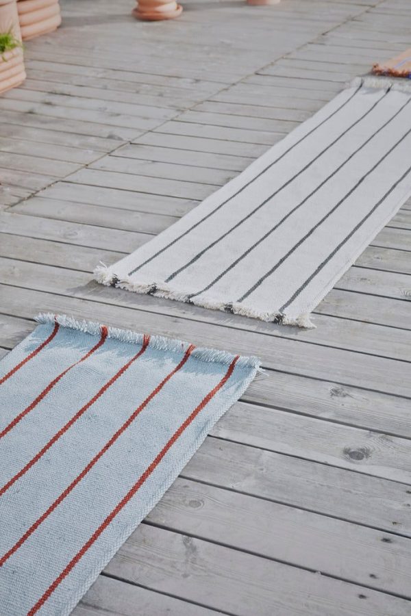 To stribede tæpper på et træterrasse afspejler Tomorrows design-æstetik; det tættere tæppe er lyseblåt med tynde røde striber, mens det andet er gråt med lyse striber, begge med kvastkanter.