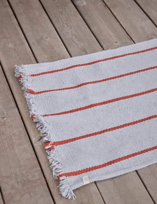 Et gråt vævet tæppe med orange vandrette striber og hvide frynser, lagt på et trægulv, eksemplificerer Tomorrows design-æstetik.
