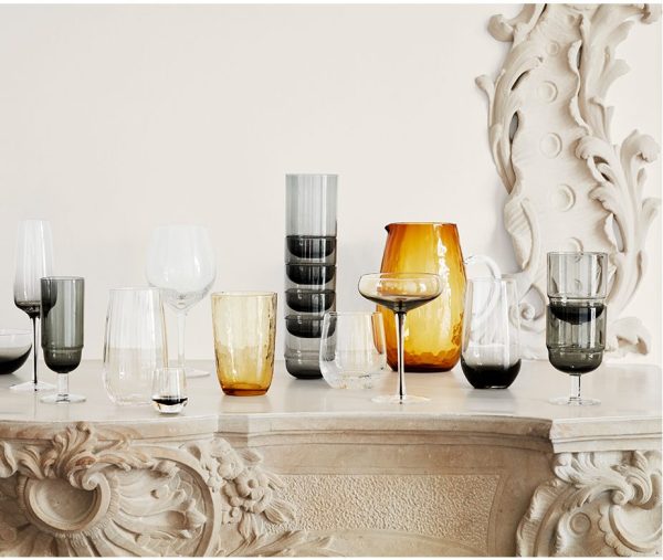 En samling af forskellige elegante BROSTE COPENHAGEN - 'Nordic Bistro' Champagneglas og vaser vist på en klassisk sten kaminhylde, med forskellige former og farver, herunder rav, sort og klart glas.