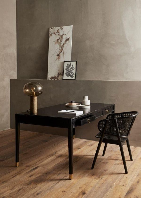 Et elegant, moderne arbejdsområde med et sort skrivebord med en gylden lampe, bøger og dekorative genstande fra TomorrowsDesign, en sort vævet stol og indrammet kunstværk lænet op ad en tekstureret grå væg på et trægulv.