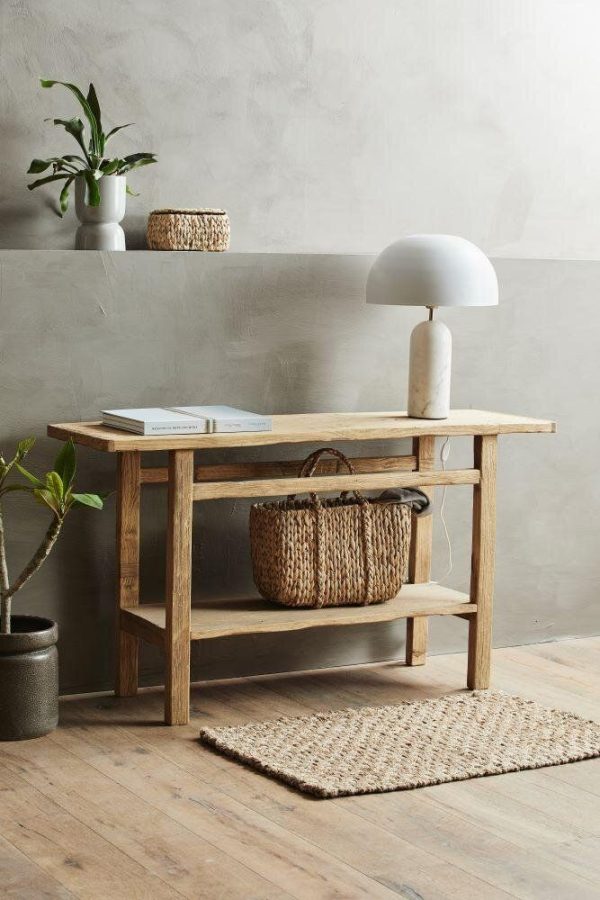 Et minimalistisk interiør med et konsolbord af træ med en hvid svampeformet lampe, en vævet kurv og en plante i en tekstureret potte. En lille bog og et vævet tæppe fuldender de fredfyldte omgivelser.