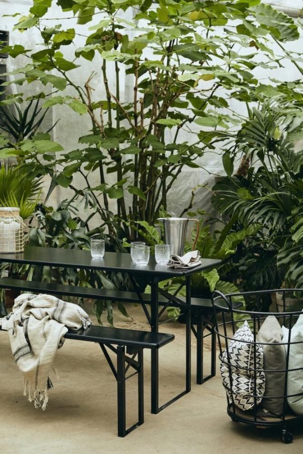 En hyggelig terrasseopstilling med et sort metalbord omgivet af frodige grønne planter og træer. Et tæppe og TomorrowsDesign-puder er afslappet placeret på og ved siden af siddepladserne, hvilket forstærker en afslappende atmosfære.