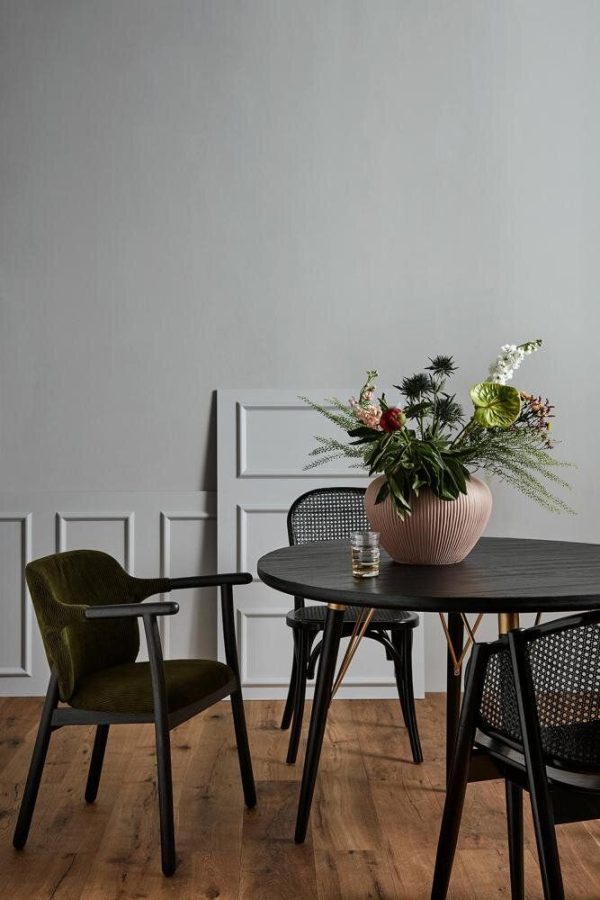 En minimalistisk spiseplads med et sort cirkulært bord og to stole, den ene betrukket med grønt. En stor lyserød krukke med en frodig buket blomster står på bordet, mod en neutral grå væg med hvidt panel. Denne indstilling byder på eksklusive designmøbler, der forstærker den æstetiske appel.