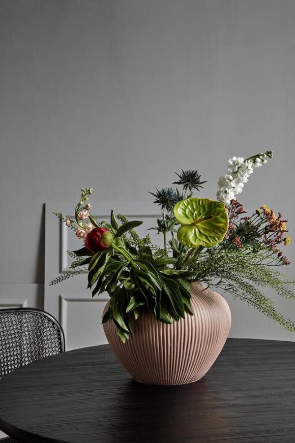 Et levende blomsterarrangement i en ribbet pink vase på et mørkt træbord med en blanding af grønne blade, hvide blomster og røde accenter mod en grå væg fra TomorrowsDesign.