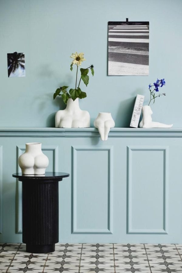 Et fredfyldt rum med en lyseblå væg med dekorative lister og et mønstret gulv. Et sort piedestalbord rummer en hvid vase, suppleret med hvide skulpturelle vaser på en hylde ovenover prydet med blomster og minimalistisk designkunst.