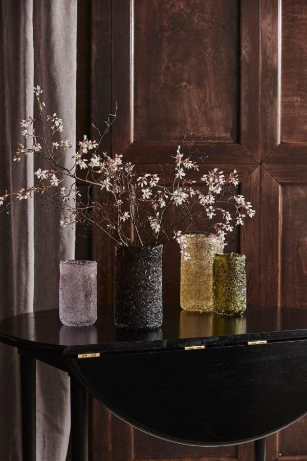 En elegant interiørscene, der viser et sort rundt bord med fire teksturerede glasvaser i forskellige farver, der hver har sarte hvide blomster. Baggrunden byder på rige, mørke træpaneler og blødt draperi fremhævet af sofistikerede designmøbler.