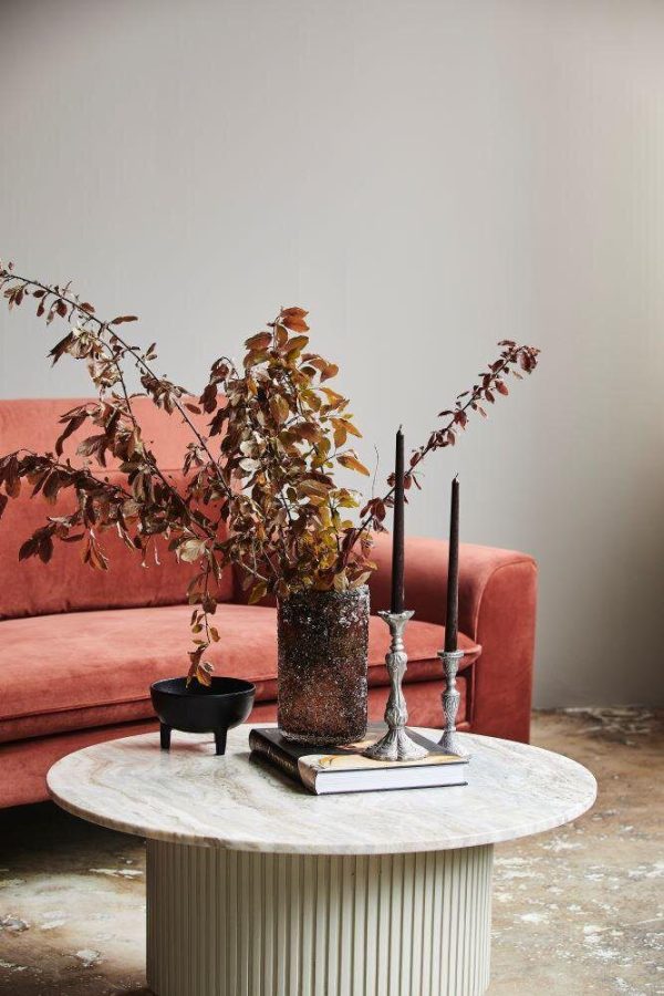 Et smart stuedesign med en koralrosa sofa, et rundt sofabord i hvid marmor prydet med bøger, en sort skål, en funklende vase med tørrede grene og to høje lysestager.
