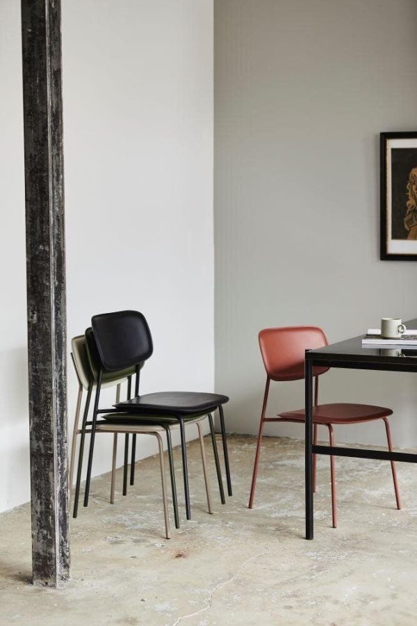Et minimalistisk interiør med en stak sorte stole, en solitær rød stol og et sort bord på et marmorgulv ved siden af en rustik søjle med neutrale vægge og Designmøbler-kunstværk synligt i baggrunden.