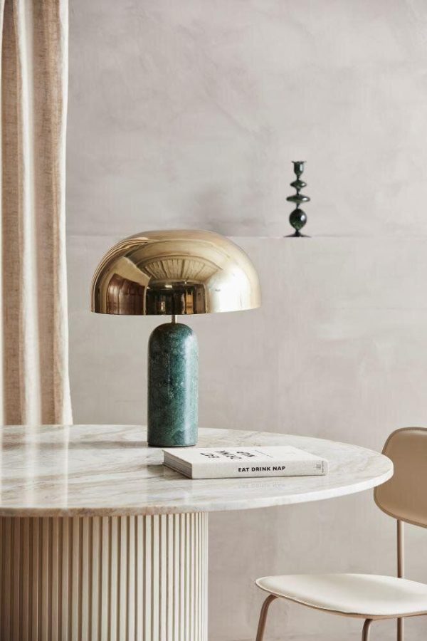 Et stilfuldt interiør med et marmorbord med en moderne metallisk lampe og en bog med titlen "eat drink nap." Bløde pastelvægge og et fløjlsgardin supplerer det minimalistiske design.