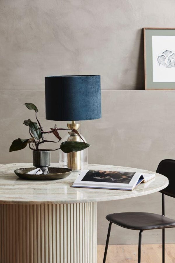 Et stilfuldt hjemmekontorhjørne med en tekstureret cremevæg, med et rundt bord med bøger og en plante, en mørk designmøblerstol og en elegant lampe med en marineblå skærm. Et indrammet billede hænger i baggrunden.