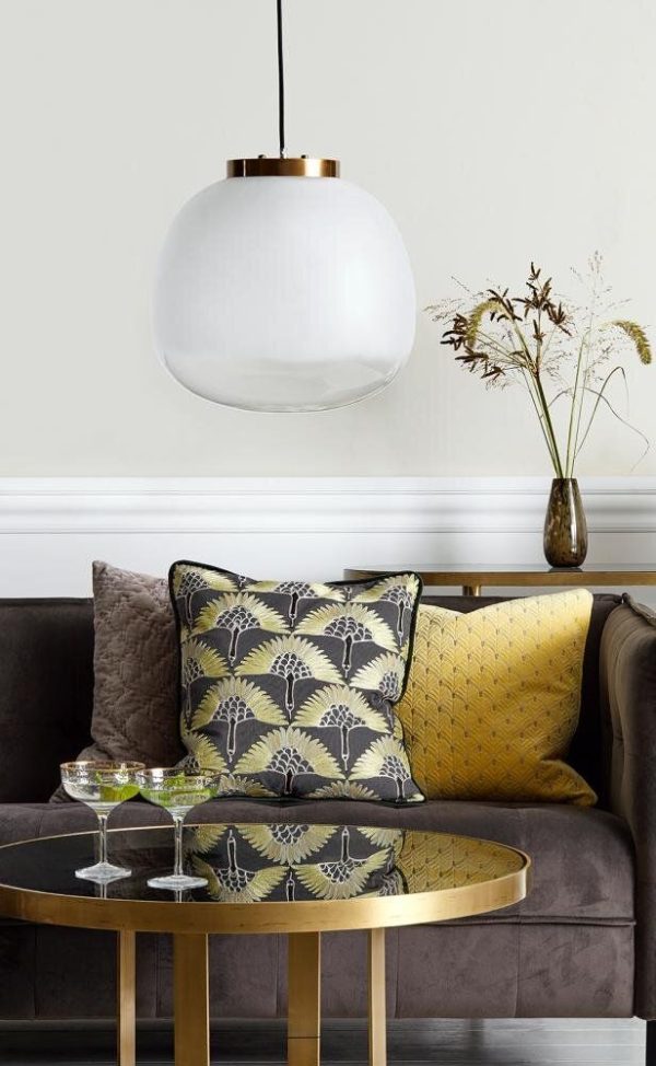 Et stilfuldt stuehjørne med en mørkegrå sofa med teksturerede hynder i forskellige mønstre og farver, et rundt guld sofabord med glas på toppen og en elegant hvid pendel overhead fra TomorrowsDesign.