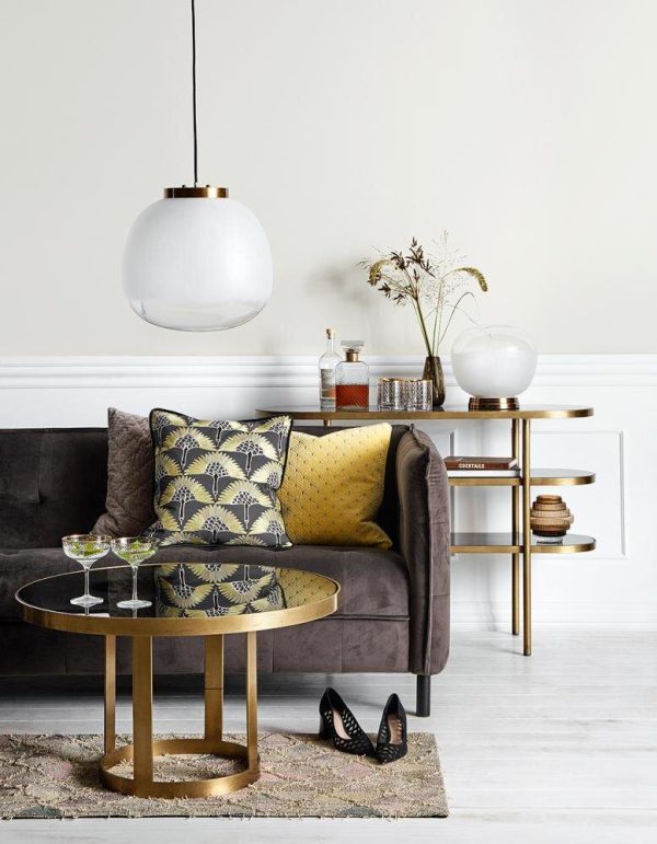Et stilfuldt stuehjørne med en mørkegrå sofa prydet med en gul mønstret pude, et guld cirkulært sofabord fra TomorrowsDesign, sidebord i glas med indretningsartikler og en stor hvid pendel. Et par sorte høje hæle sidder på gulvet.