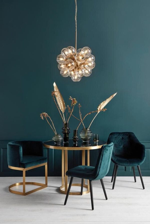 En elegant spisestue med en mørk blågrøn væg med en boblelysekrone af guld og glas hængende over et cirkulært bord flankeret af plys-fløjlsstole med guldaccenter. Stilfulde vaser og dekorative fjer fuldender det sofistikerede design.