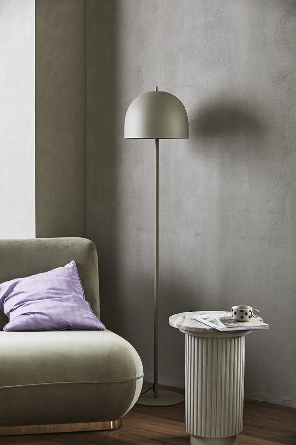 Et minimalistisk interiørdesign i stuehjørnet med en grøn lænestol med en lilla pude, et hvidt cylindrisk sidebord med bøger og en kop og en elegant gulvlampe med en stor kuppelskærm.
