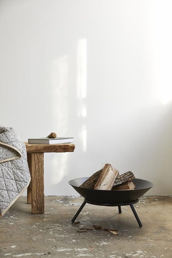 Et minimalistisk interiør med et simpelt sidebord med en træplade og et tæppe draperet over, ved siden af en sort metalskållignende bålplads med brænde på et betongulv. Lyset strømmer gennem et vindue op på væggen.