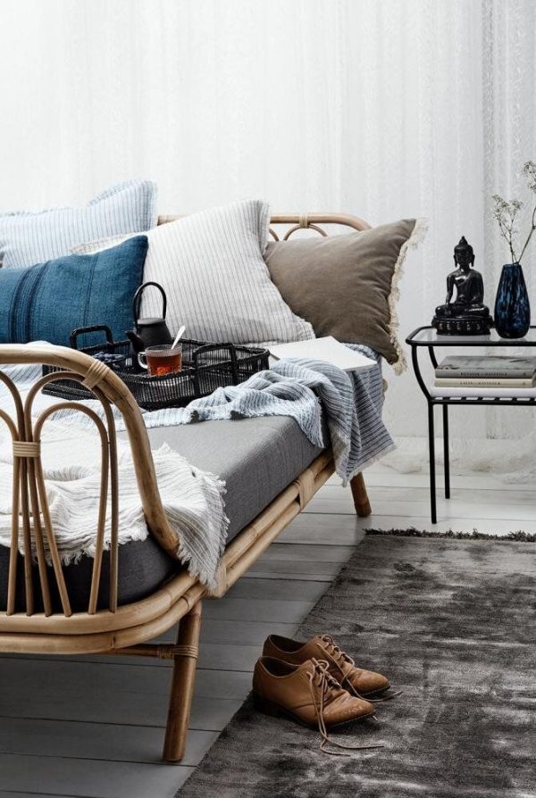 Et hyggeligt stuehjørne med en rattansofa prydet med blå og grå puder, et hvidt tæppe og et lille sidebord med tekande. Brune designmøbler sko er placeret pænt på et gråt tæppe.