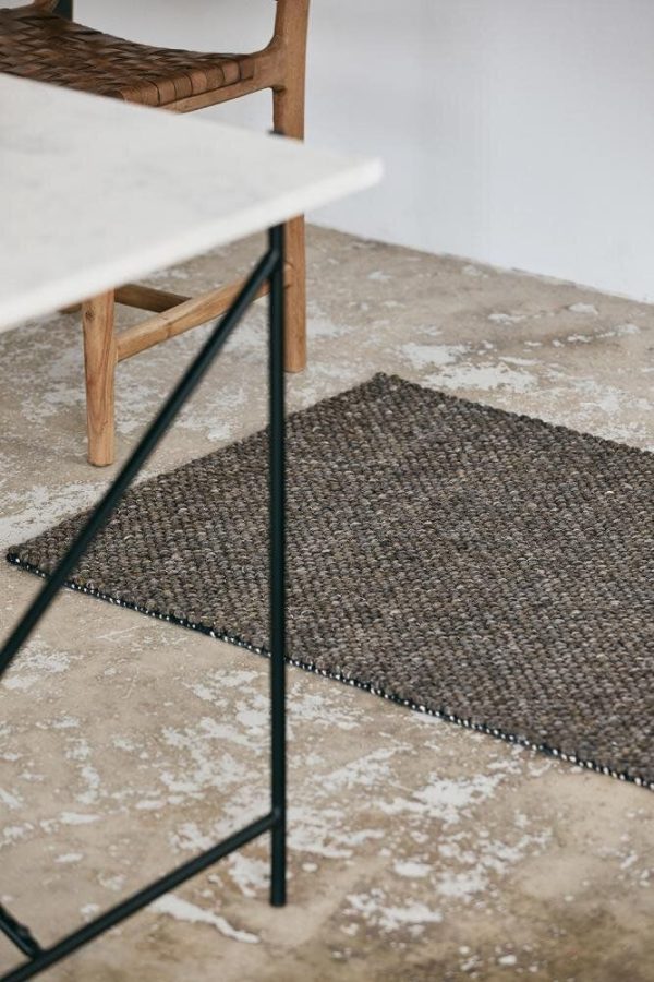 Et minimalistisk indvendigt rum med et gråspættet betongulv med et tekstureret vævet tæppe, sat ved siden af et moderne bord med sorte metalben og en delvist synlig træ-spisestuestol, kurateret af TomorrowsDesign.