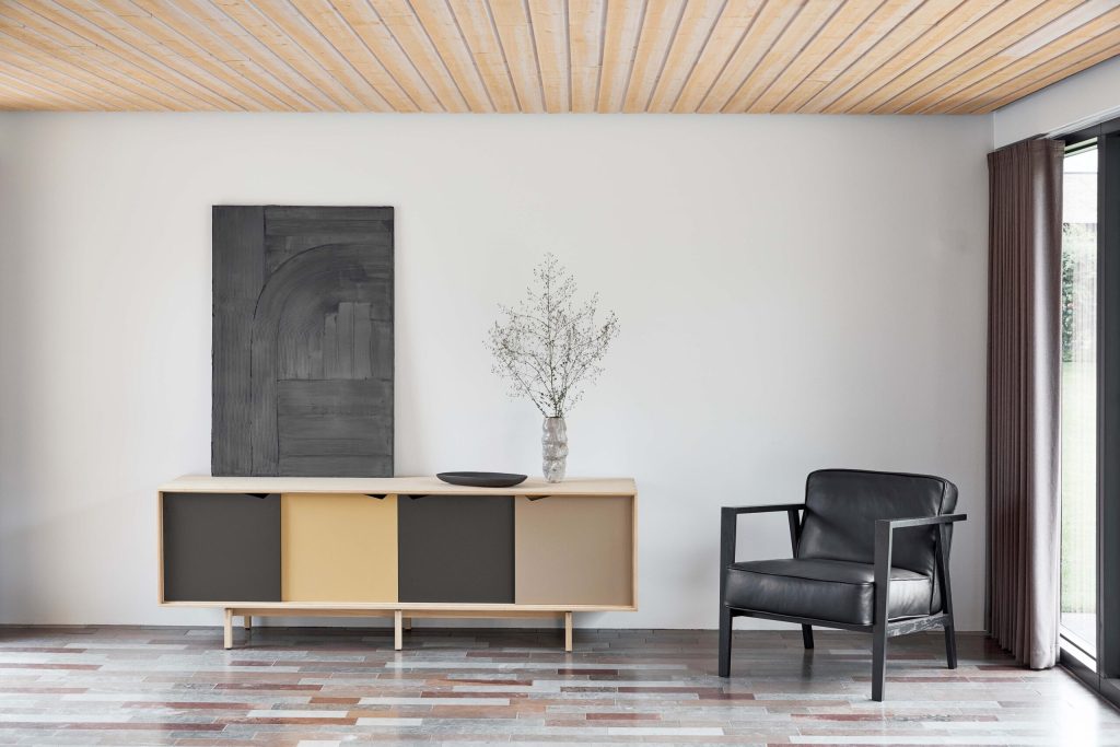 Et minimalistisk værelse med et flerfarvet klinkegulv, med en elegant skænk med beige og gule paneler, toppet med en vase og et stort abstrakt maleri over det. en sort lænestol sidder til højre ved en glasdør.